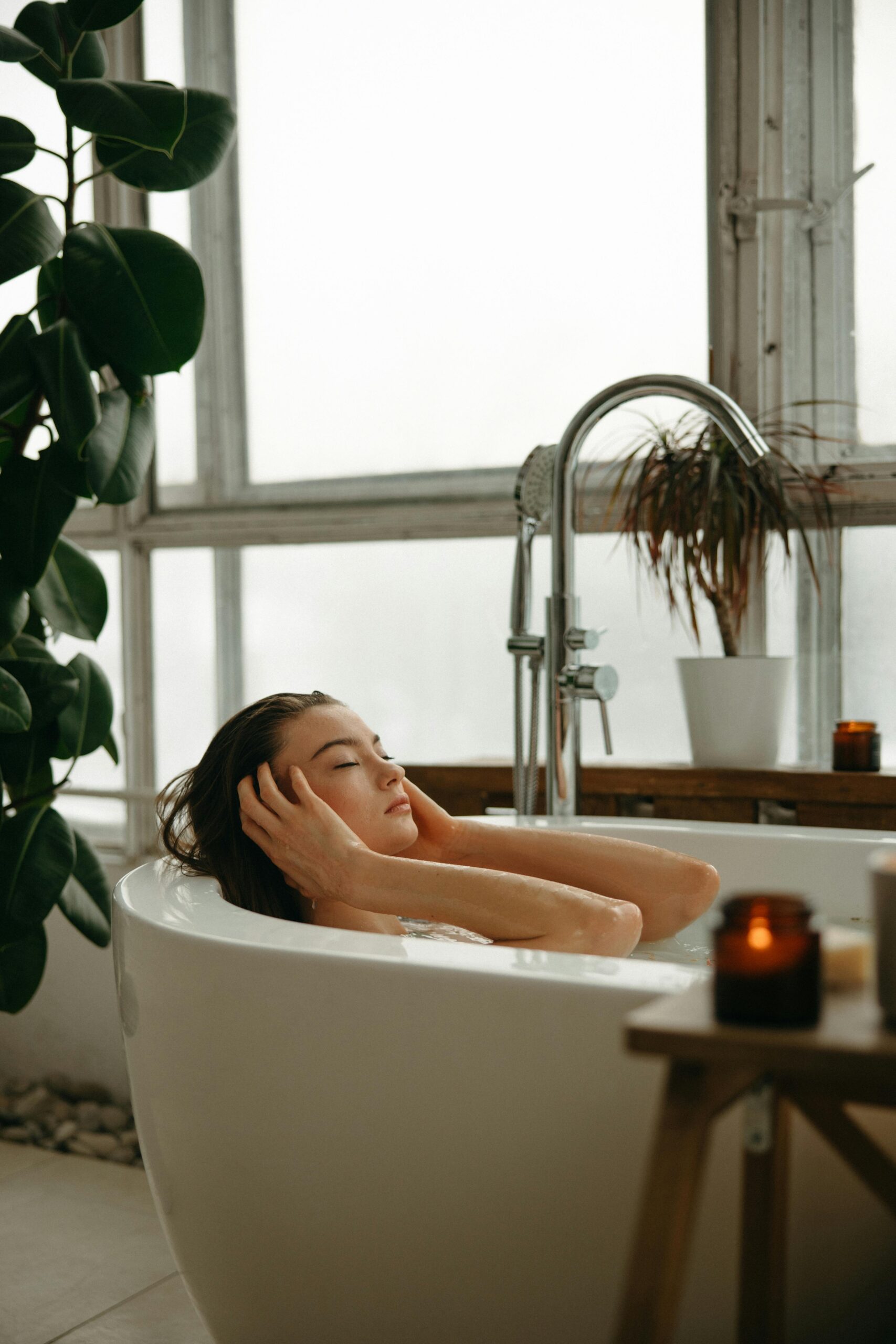 A woman enjoys a serene bath in a light-filled bathroom with plants and candles.